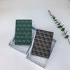 A dark green geometric print passport wallet displayed in a clear presentation box on a white surface.