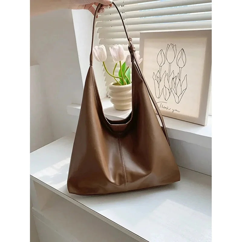 A brown PU leather slouchy hobo shoulder bag hanging on a hanger in front of a window.