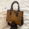 A brown patterned ladies' shoulder tote bag resting on a white blanket with a card nearby.