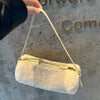 A close-up shot of a cream-colored, fluffy cylinder shoulder bag with a gold zipper and white strap hanging against a gray te