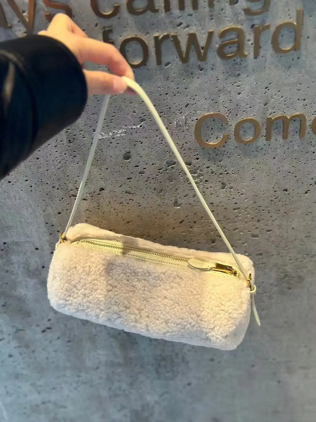 A close-up shot of a cream-colored, fluffy cylinder shoulder bag with a gold zipper and white strap hanging against a gray te