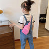 A young girl wearing denim overalls and a pink nylon crossbody bag stands next to a wooden cabinet.