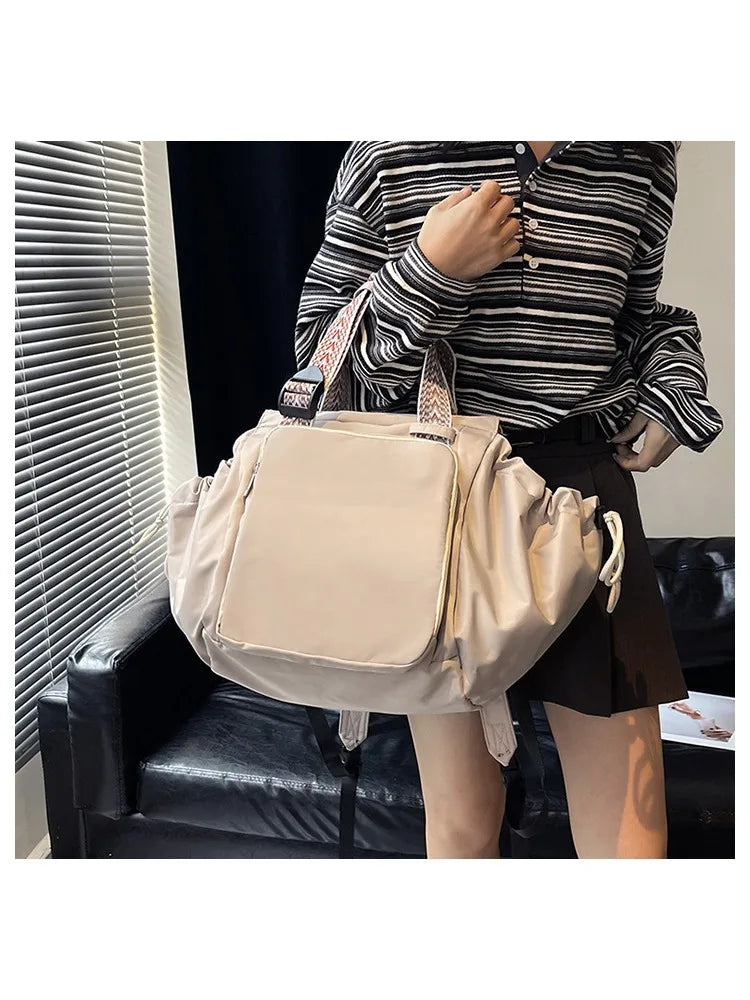 A woman holds a beige nylon shoulder bag with a patterned strap in an indoor setting.