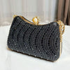 A black rhinestone evening clutch with gold accents and a pearl closure resting on a white surface.