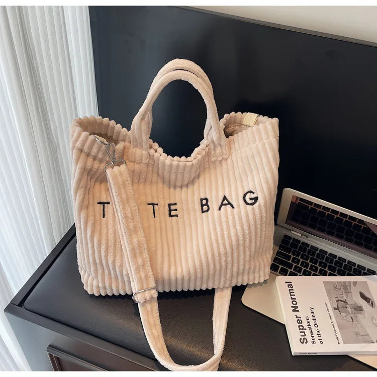 Sac à épaule beige côtelé avec une sangle en cuir, exposé sur une surface sombre à côté d'un ordinateur portable.