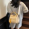 A woman is wearing a beige nylon crossbody bag with an orange accent on her gray shirt, standing in front of dark stairs.