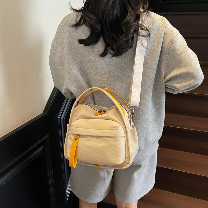 A woman is wearing a beige nylon crossbody bag with an orange accent on her gray shirt, standing in front of dark stairs.
