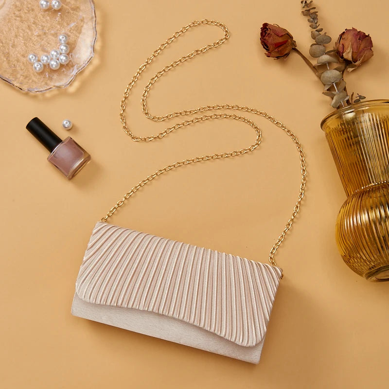 A beige satin evening clutch with a gold chain strap is displayed next to a gold chain and a small cosmetic product.