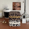 A stack of leopard print PU leather jewelry storage boxes next to a silver perfume bottle and a hand wearing rings.