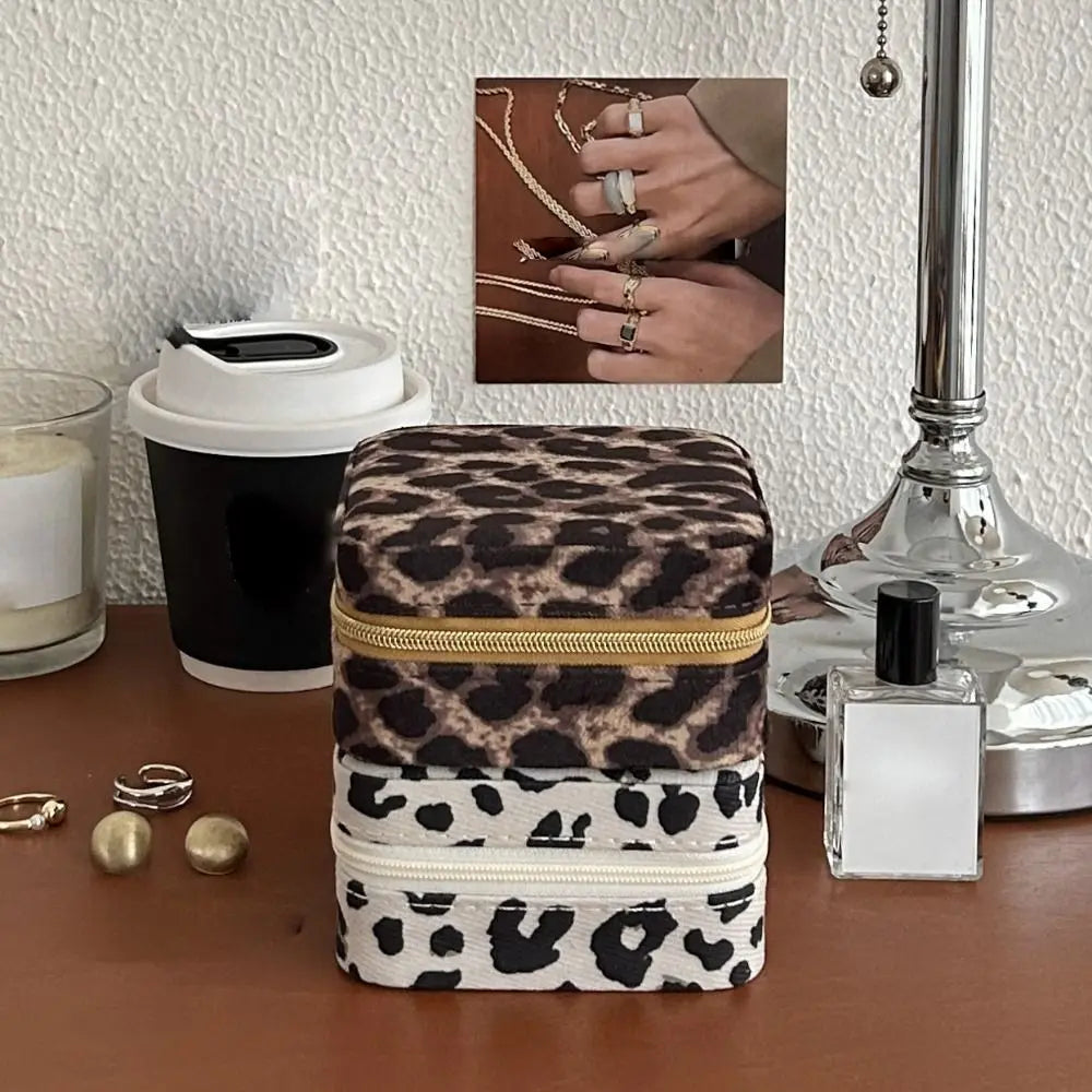 A stack of leopard print PU leather jewelry storage boxes next to a silver perfume bottle and a hand wearing rings.