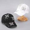 A black mesh snapback baseball cap decorated with silver floral embroidery, displayed next to a white version on a wooden bas