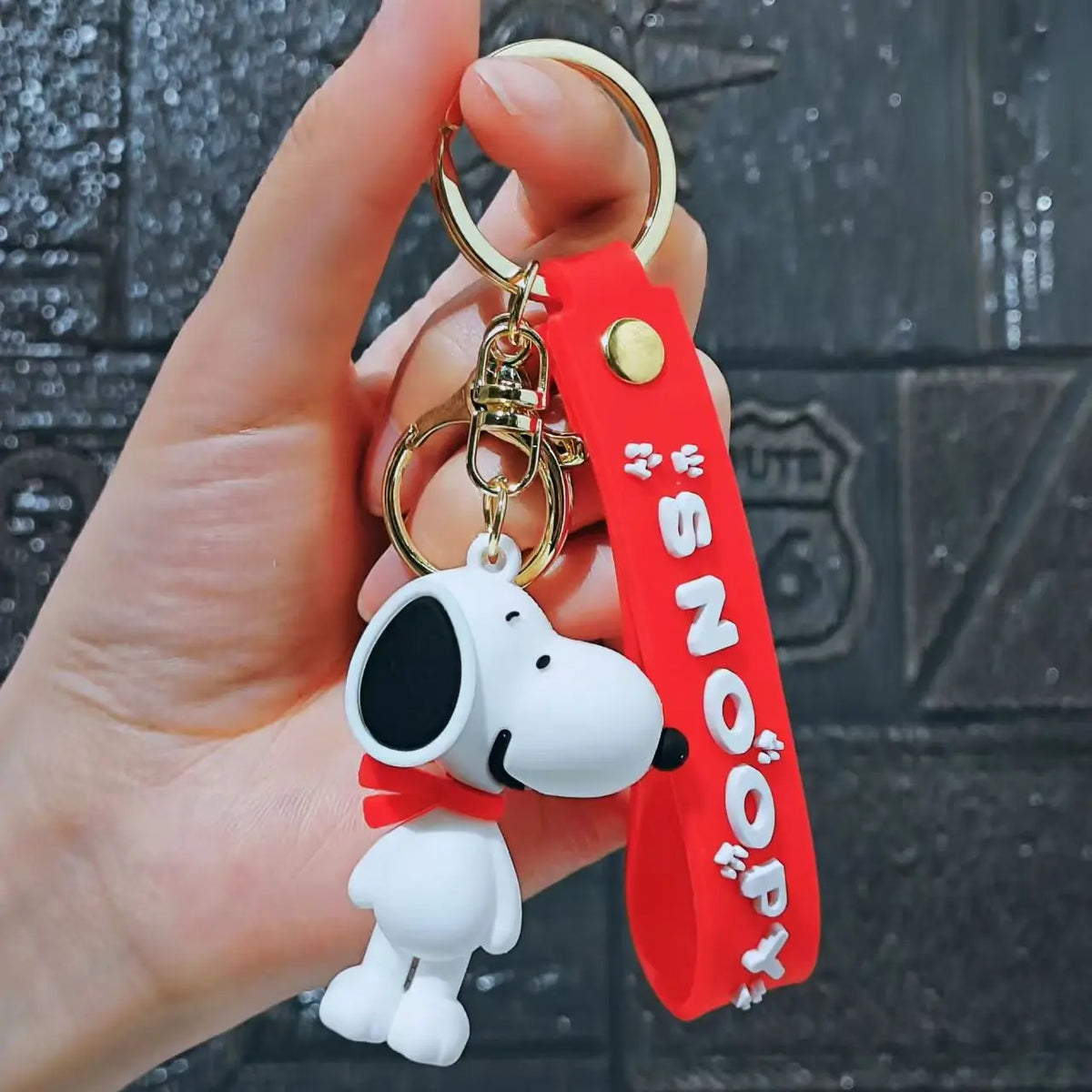 A hand holds a red silicone keychain featuring a white Snoopy face and gold hardware.