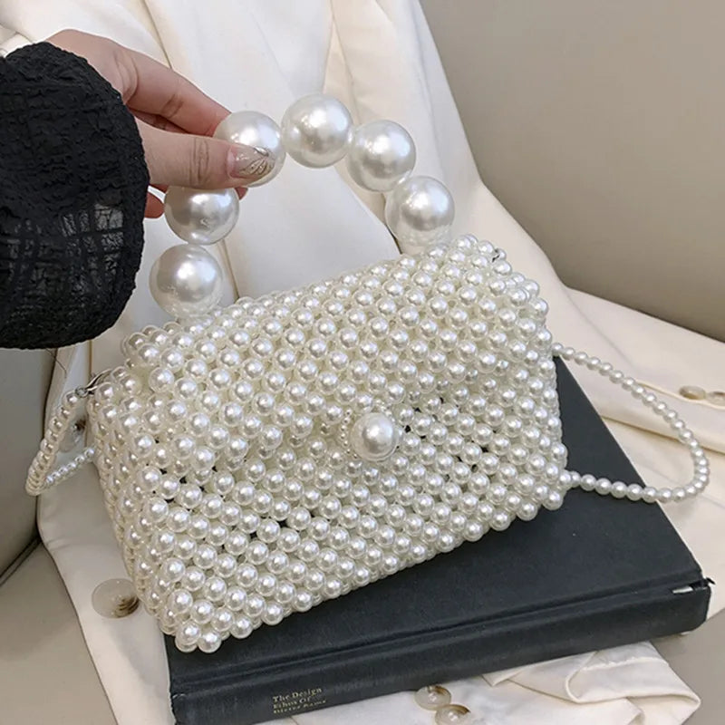 A handmade pearl beaded clutch bag with a strap resting on a dark book, next to a hand showing several pearls.