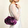 A woman wearing a white dress holds a deep purple rose floral evening clutch bag with a silver chain strap.