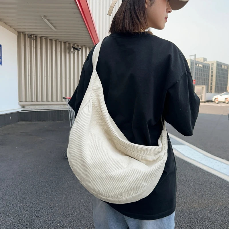 A person wearing a black shirt and light blue jeans is carrying a cream-colored corduroy casual crossbody shoulder bag.