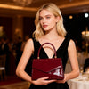 A blonde woman holds a glossy burgundy evening handbag in a black dress at an event.