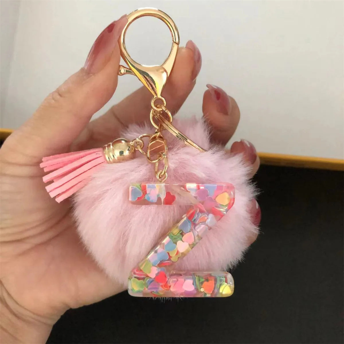 A hand holds a custom initial letter keychain featuring a shiny resin pendant with a pink alphabet charm and pink feathers.