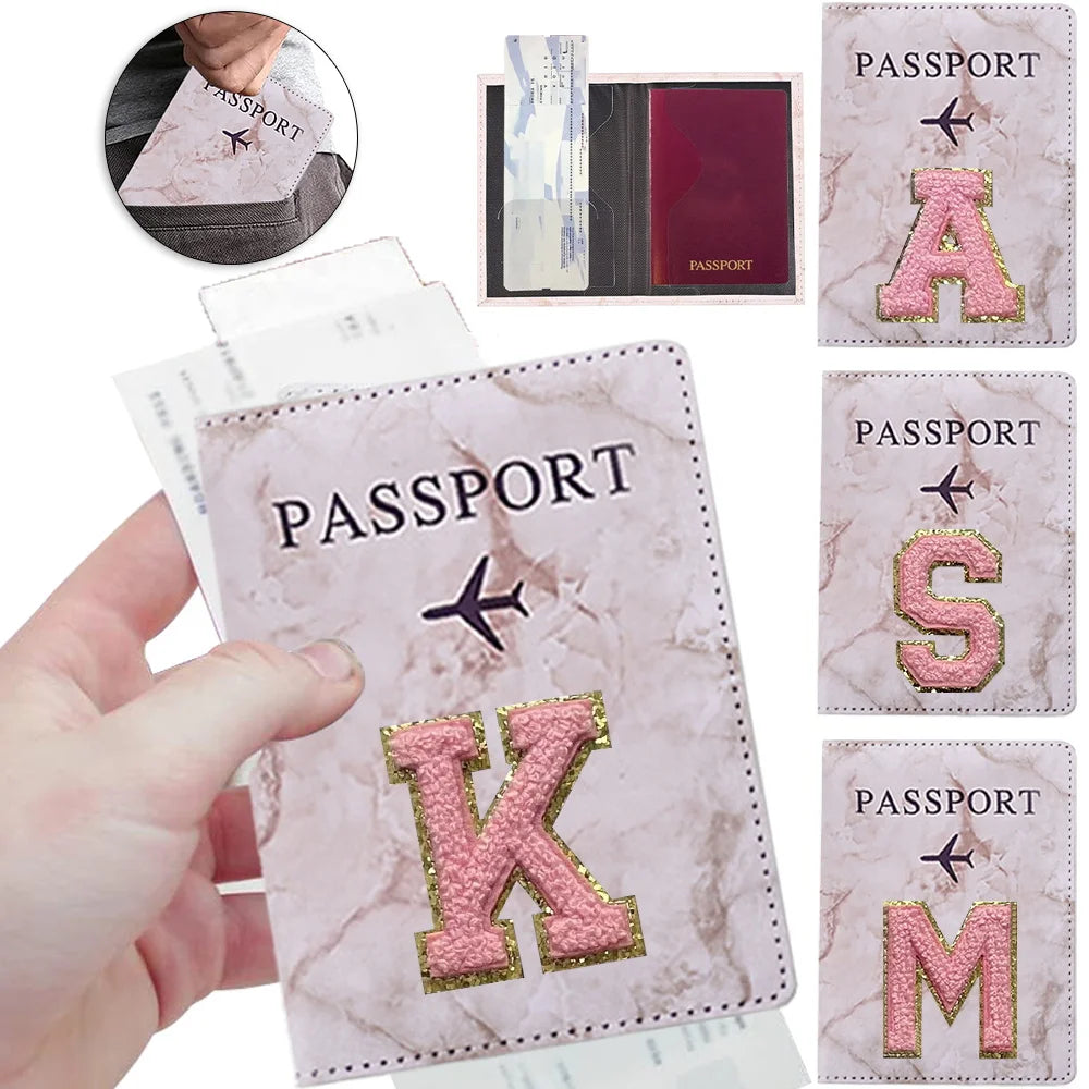 Una mano sostiene una funda de pasaporte con patrón de mármol rosa y una inicial K dorada, con otras fundas visibles.