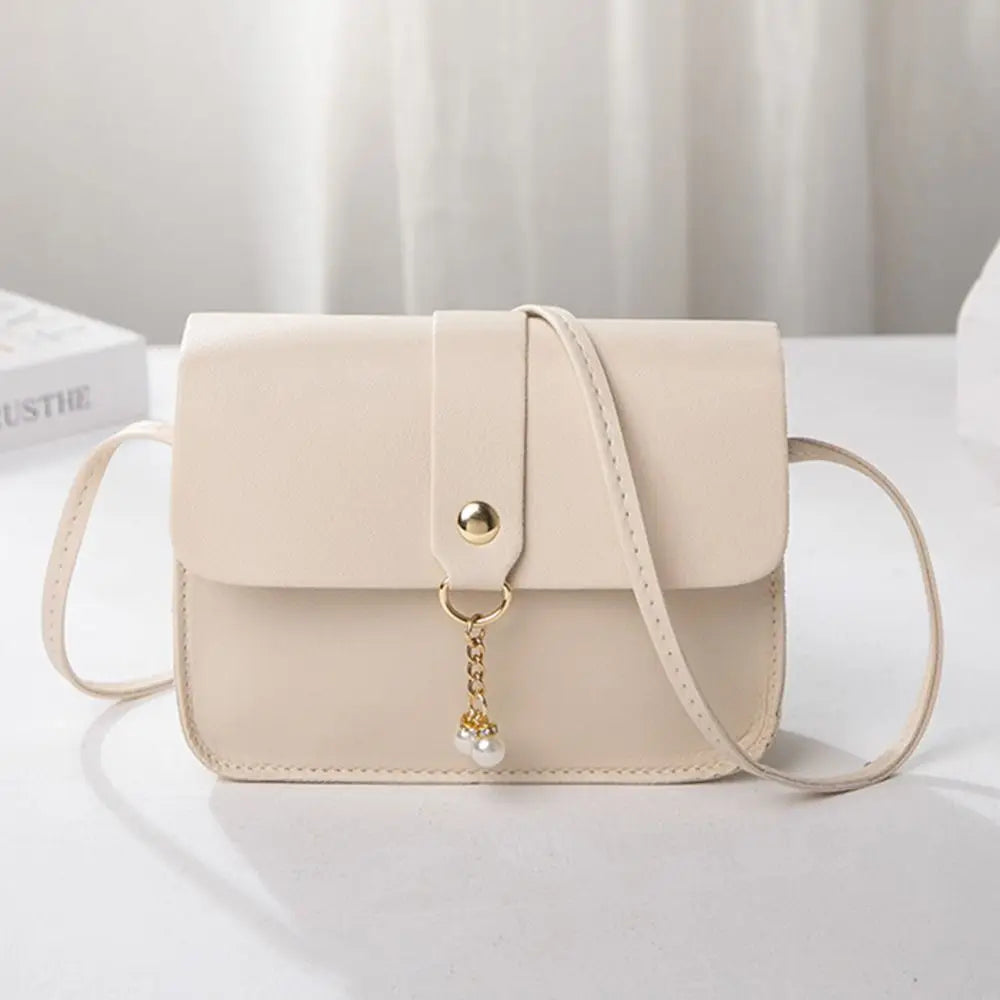 A beige fashion square crossbody bag with a gold chain charm resting on a white surface.