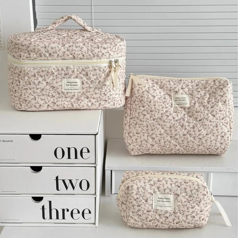 A set of three floral print quilted cosmetic bags in beige and white are displayed on a white shelf.