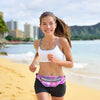 A young woman wearing a white sports top and dark shorts runs along a sandy beach near the ocean.
