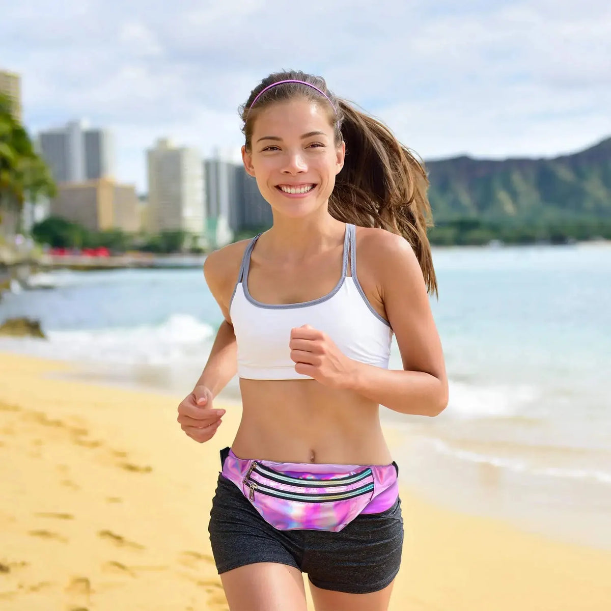 A young woman wearing a white sports top and dark shorts runs along a sandy beach near the ocean.