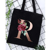 Black canvas tote bag featuring a large gold letter R with floral accents