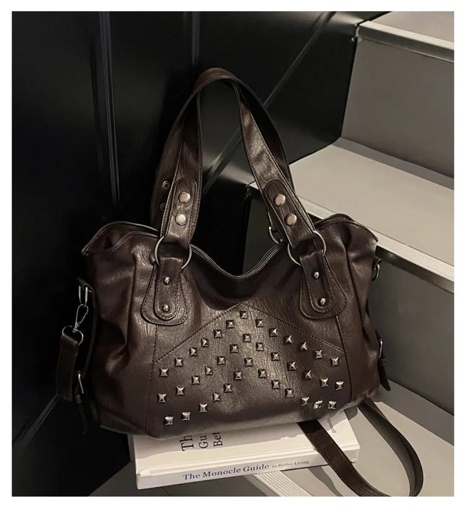 A dark brown punk rivet leather tote bag is displayed in a box next to a stack of books.