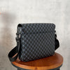 A black patterned flap crossbody bag resting on a wooden stool against a gray concrete background.