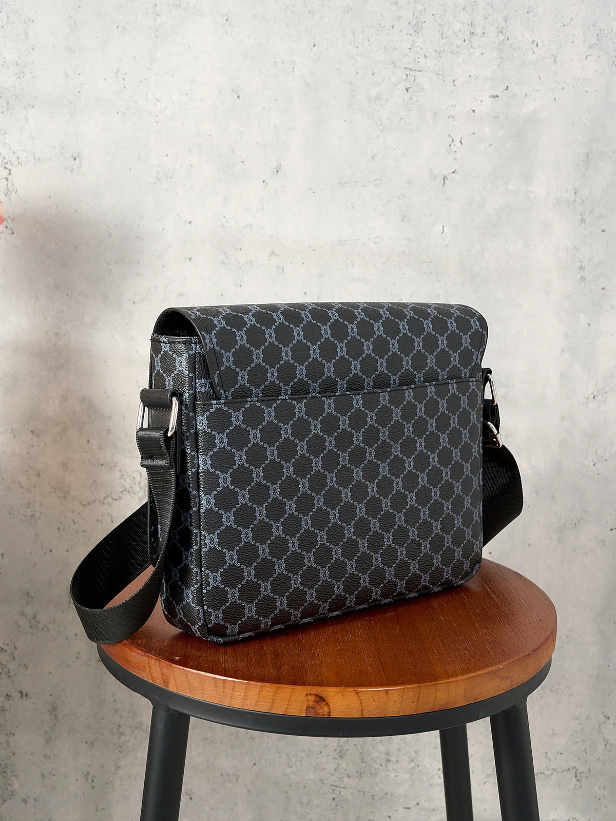 A black patterned flap crossbody bag resting on a wooden stool against a gray concrete background.