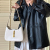 A woman holds a white leather hobo shoulder bag with a gold chain strap against a background of light blinds and a black leat