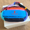 A colorful patchwork color block fanny pack with purple, orange, pink, blue, and dark blue sections resting on a white box.