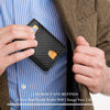 A hand holds a black slim leather wallet with a gold tag against a blue striped shirt and brown jacket.