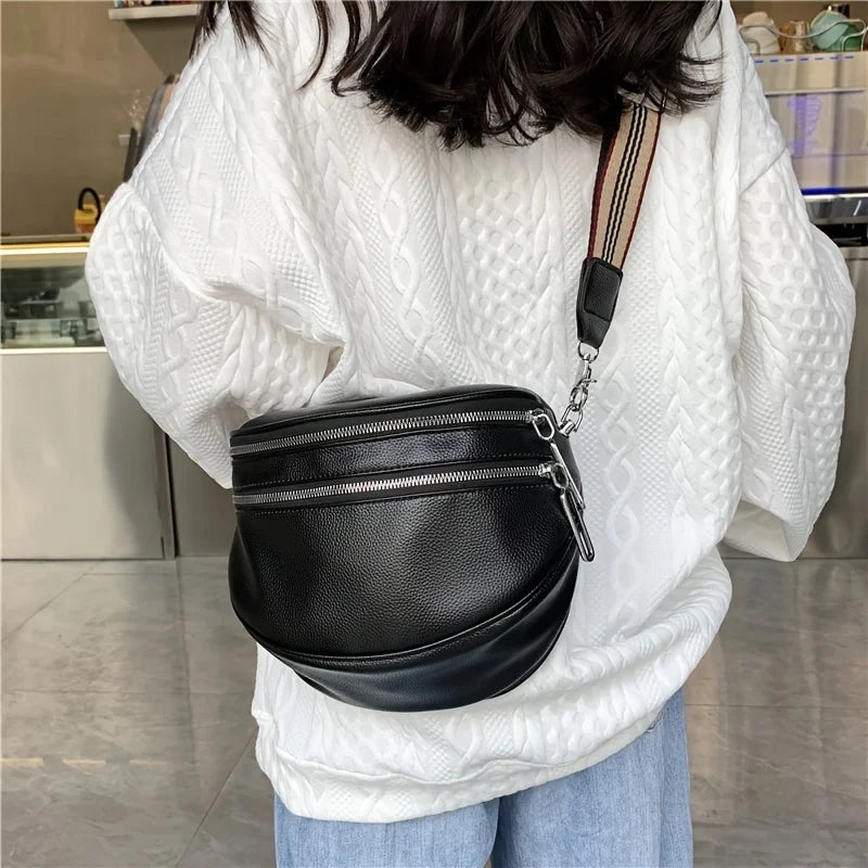 A black soft leather crossbody bag with double zippers and an adjustable strap is worn by a person in a white sweater.