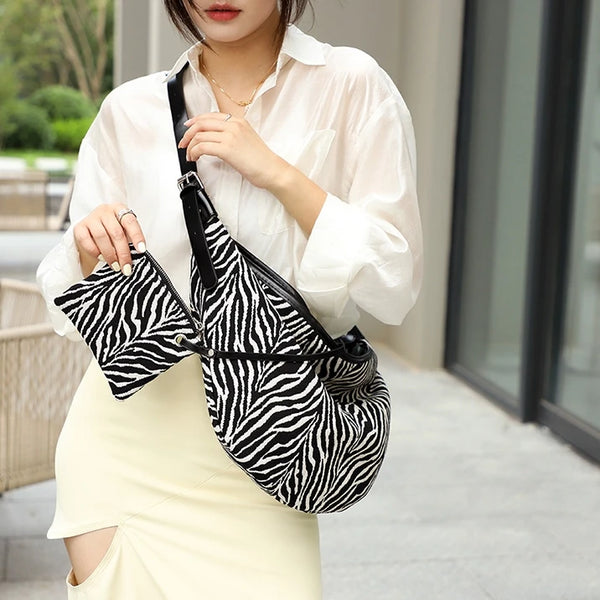 A woman wearing a white shirt and light skirt holds a black canvas zebra print shoulder crossbody bag.