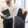 A man in a white shirt and black pants holds a black laptop sleeve while standing near a window.