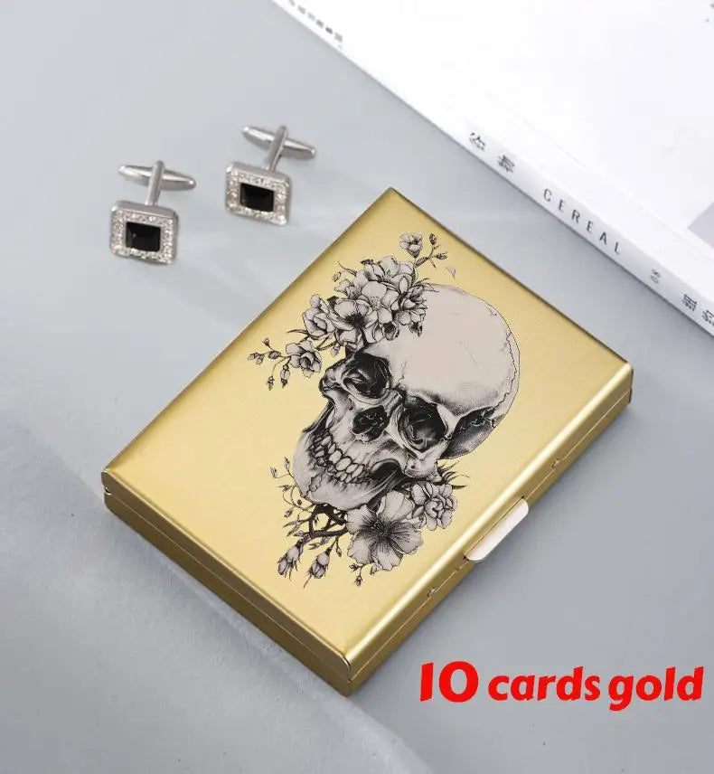 A gold card holder featuring a detailed skull design, with two silver earrings displayed nearby.
