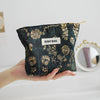 A dark teal floral canvas makeup bag with gold embroidery is held in a hand.