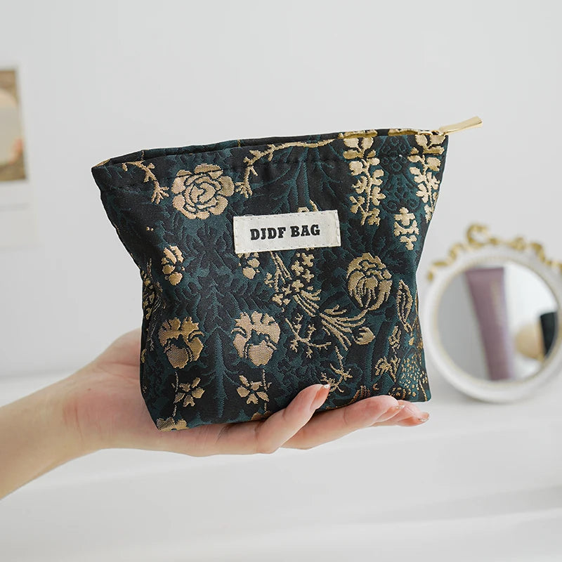 A dark teal floral canvas makeup bag with gold embroidery is held in a hand.