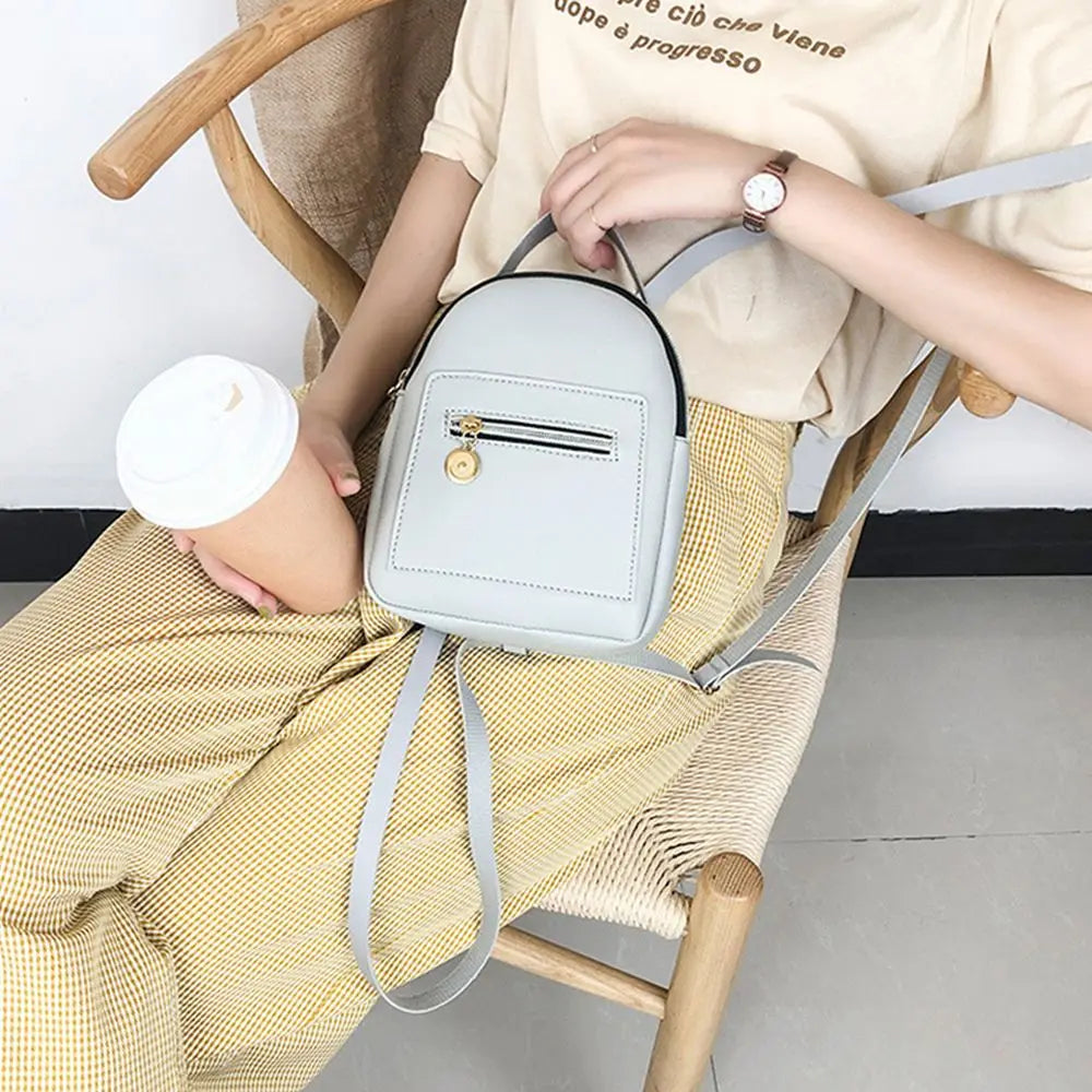 A woman is sitting in a wooden chair holding a light gray waterproof PU mini backpack with a leather strap.