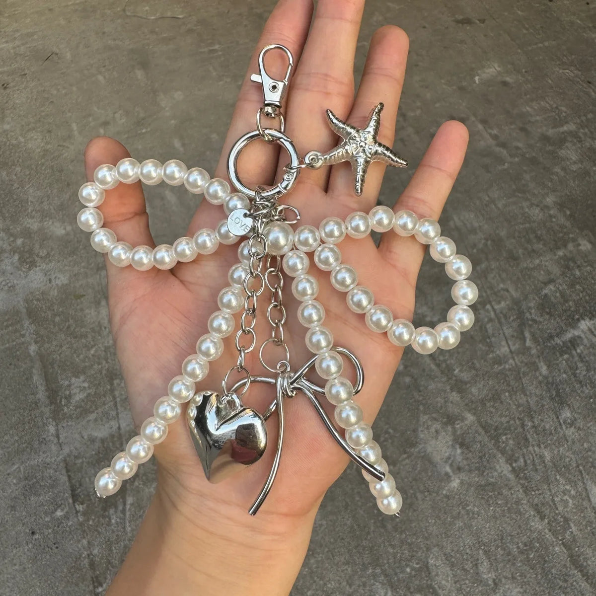 A hand holds a silver pearl bowknot keychain featuring a starfish charm and a heart pendant.