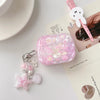 Étui d'écouteurs AirPods avec nœud de fleur rose et charms argentés sur une surface blanche.