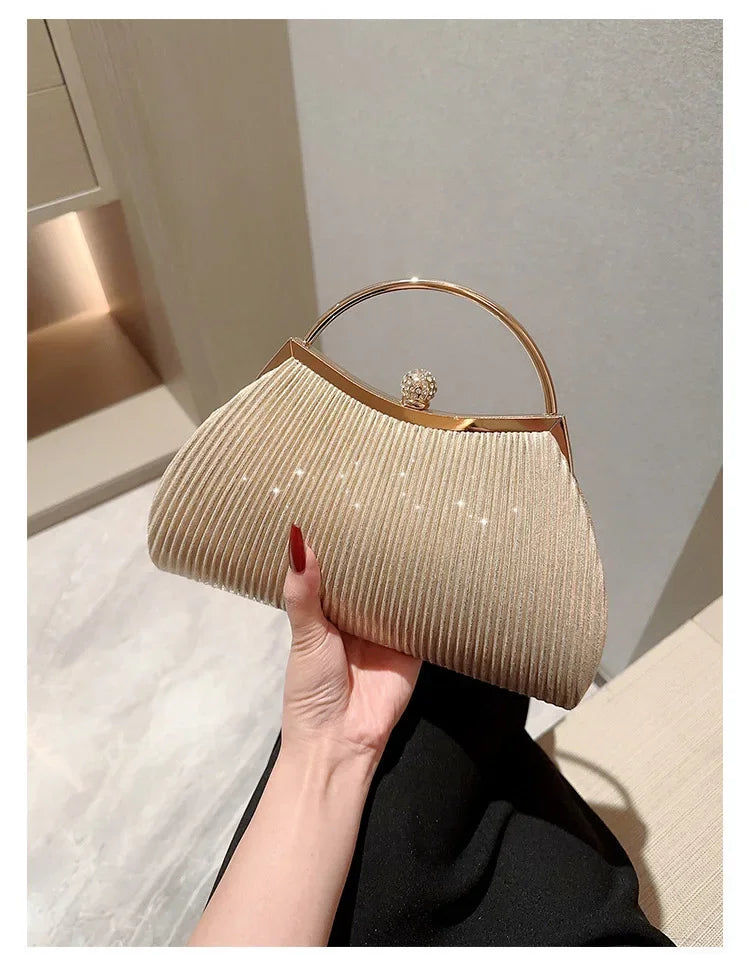 A beige pleated glitter evening handbag with a gold top handle and chain strap being held by a hand.