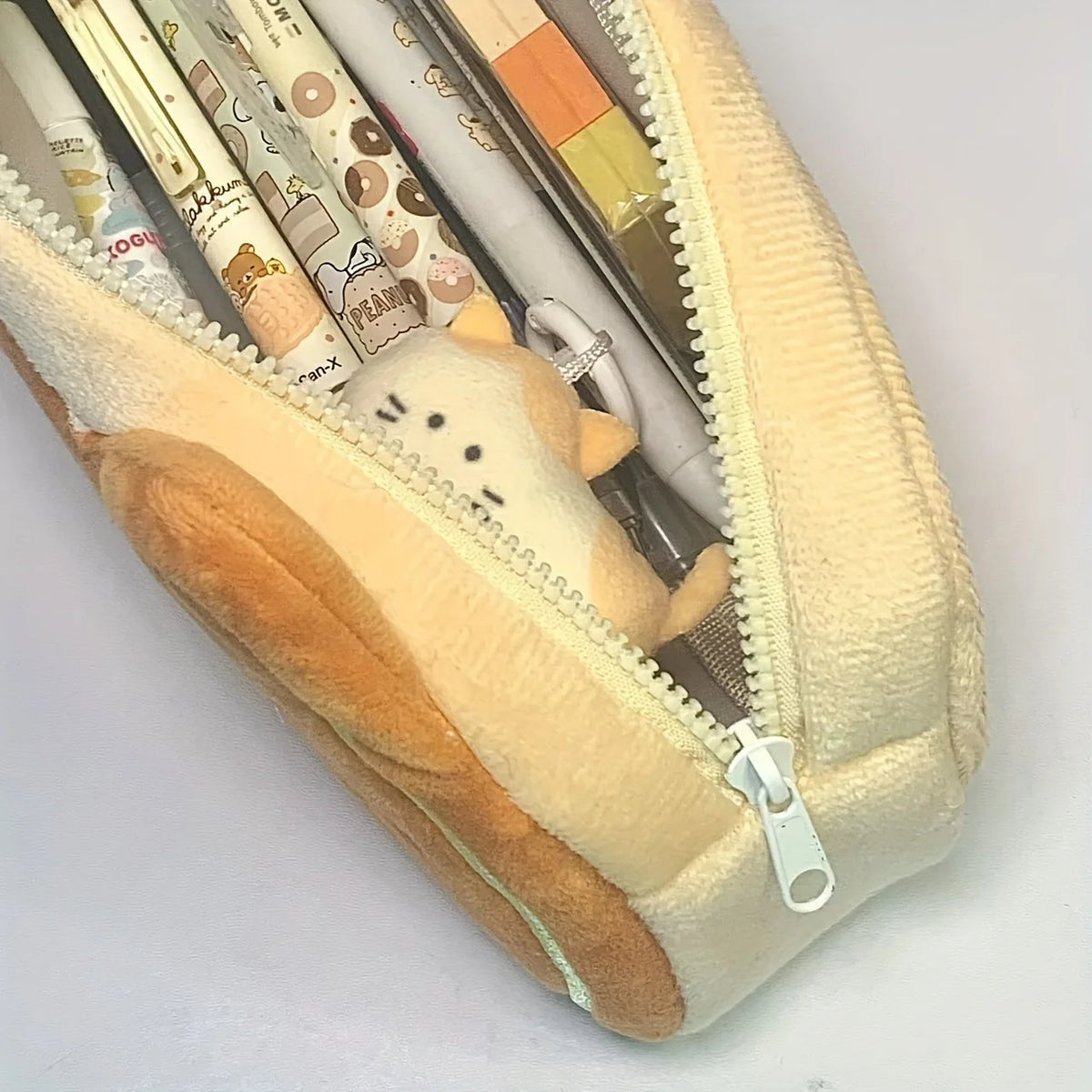 A beige pencil pouch with a zipper, featuring a caramel cat plush toy and various pencils inside.