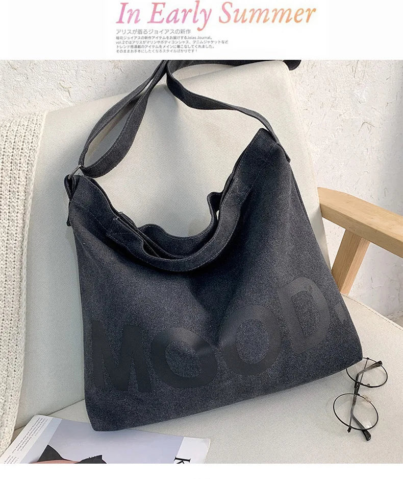 A dark gray canvas tote bag featuring a large letter design, displayed on a white surface.