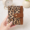 A hand holds a brown and black leopard print compact wallet with a leather strap next to a white clock.