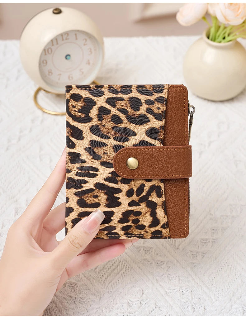 A hand holds a brown and black leopard print compact wallet with a leather strap next to a white clock.