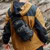 A man wearing a tan and black jacket is carrying a tactical chest bag on his back in an outdoor setting.
