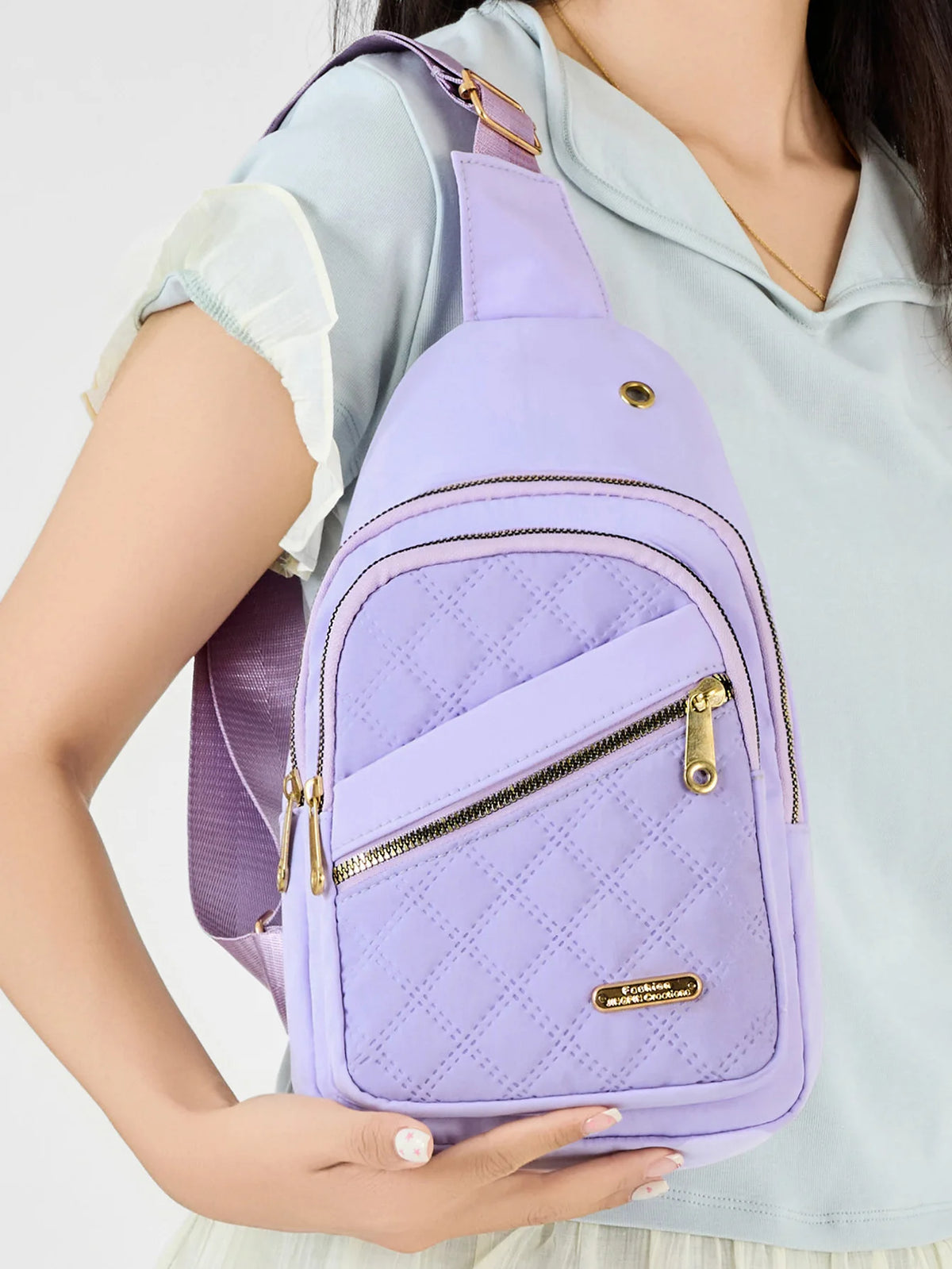 Un sac bandoulière à motif matelassé violet avec une fermeture éclair dorée porté par une personne en chemise bleu clair.