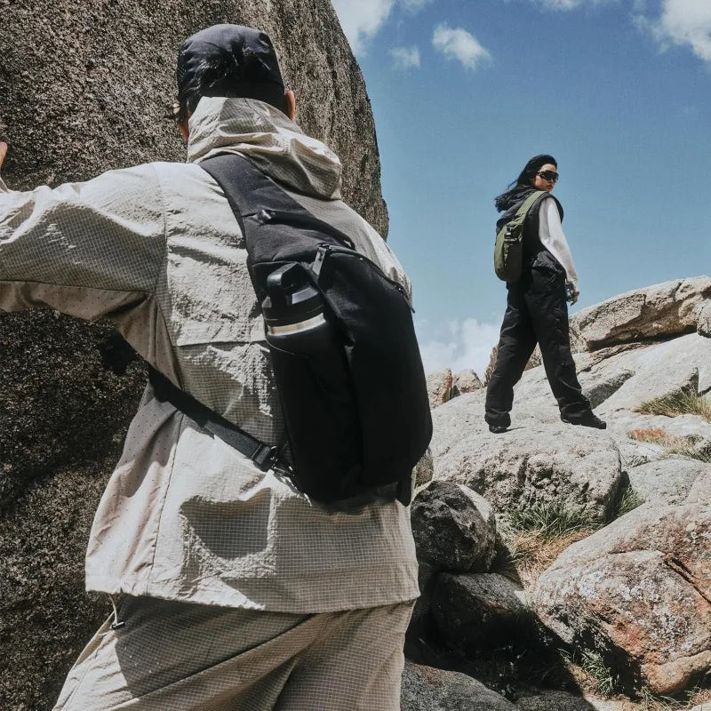 A person wearing a light jacket and carrying a black crossbody sling bag is standing on rocks under a blue sky.
